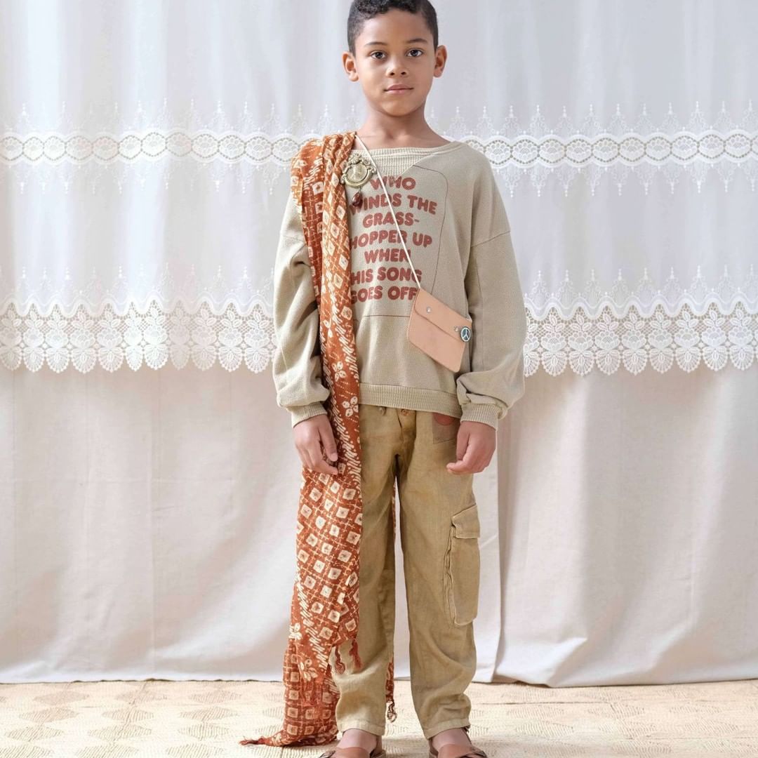 Young boy poses in a casual outfit consisting of a beige sweatshirt with text, cargo pants, and sandals against a light backdrop.