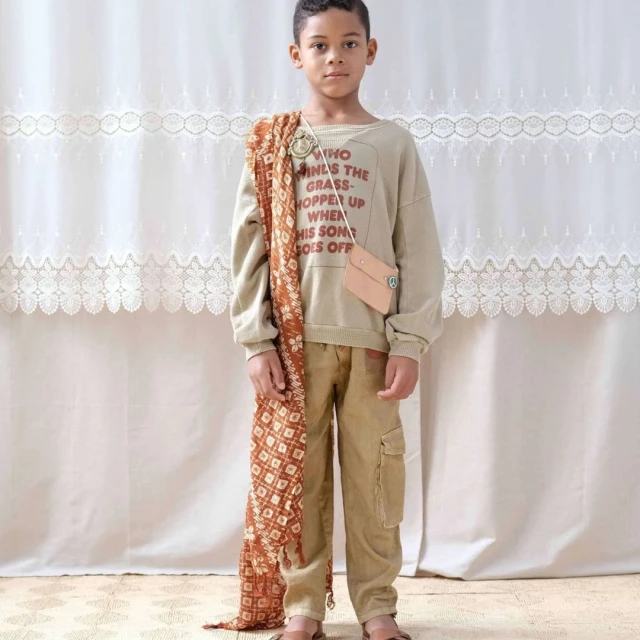 Young boy poses in a casual outfit consisting of a beige sweatshirt with text, cargo pants, and sandals against a light backdrop.