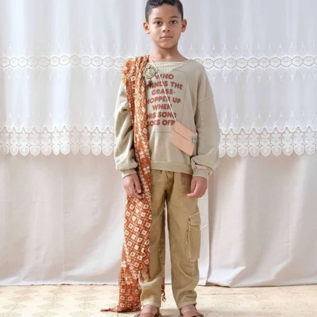 Young boy poses in a casual outfit consisting of a beige sweatshirt with text, cargo pants, and sandals against a light backdrop.