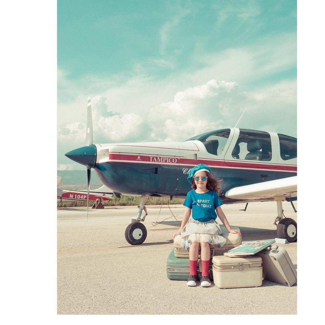 Young girl with curly hair wearing a blue beret and sitting on a stack of vintage suitcases in front of a small airplane on a tarmac.