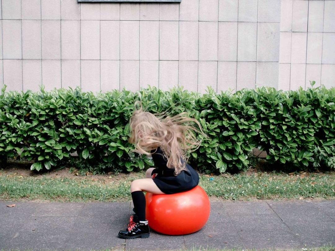 A girl with flowing blond hair sits on a large orange exercise ball in an outdoor scene with a wall and green bushes.