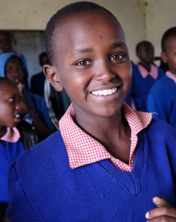 Portrait of a smiling young African student wearing a blue sweater and red-and-white checkered collar, surrounded by other students.