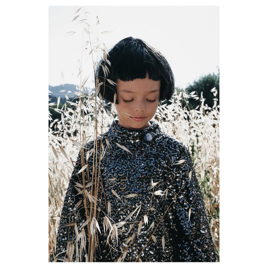 Portrait of a young child with dark hair wearing a sequined coat in a grassy field on a sunny day, creating a captivating fashion image.