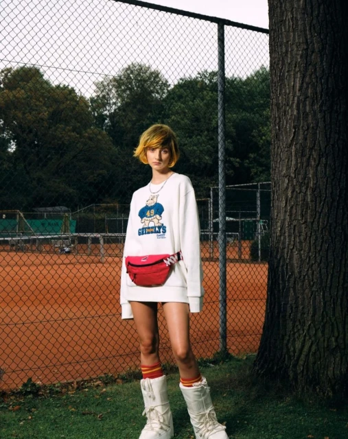 Woman posing in a sweatshirt and red belt bag in front of a tennis court fence with trees and grass in the background.
