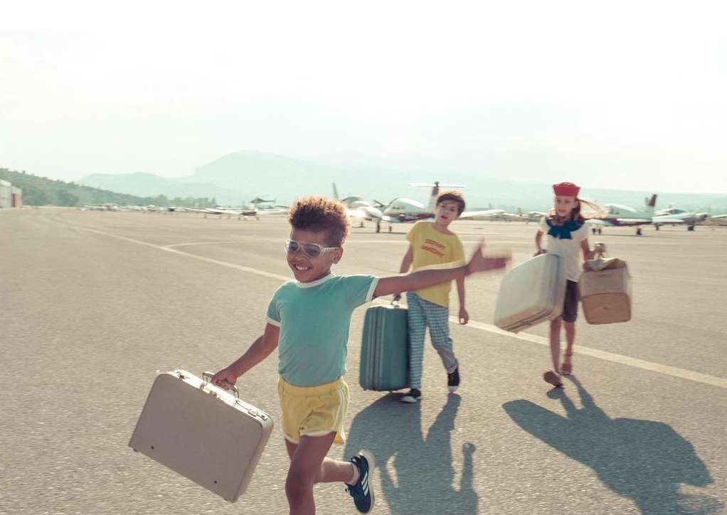 Three children run with vintage suitcases on an airport runway near small airplanes on a sunny day, creating a vintage travel scene.