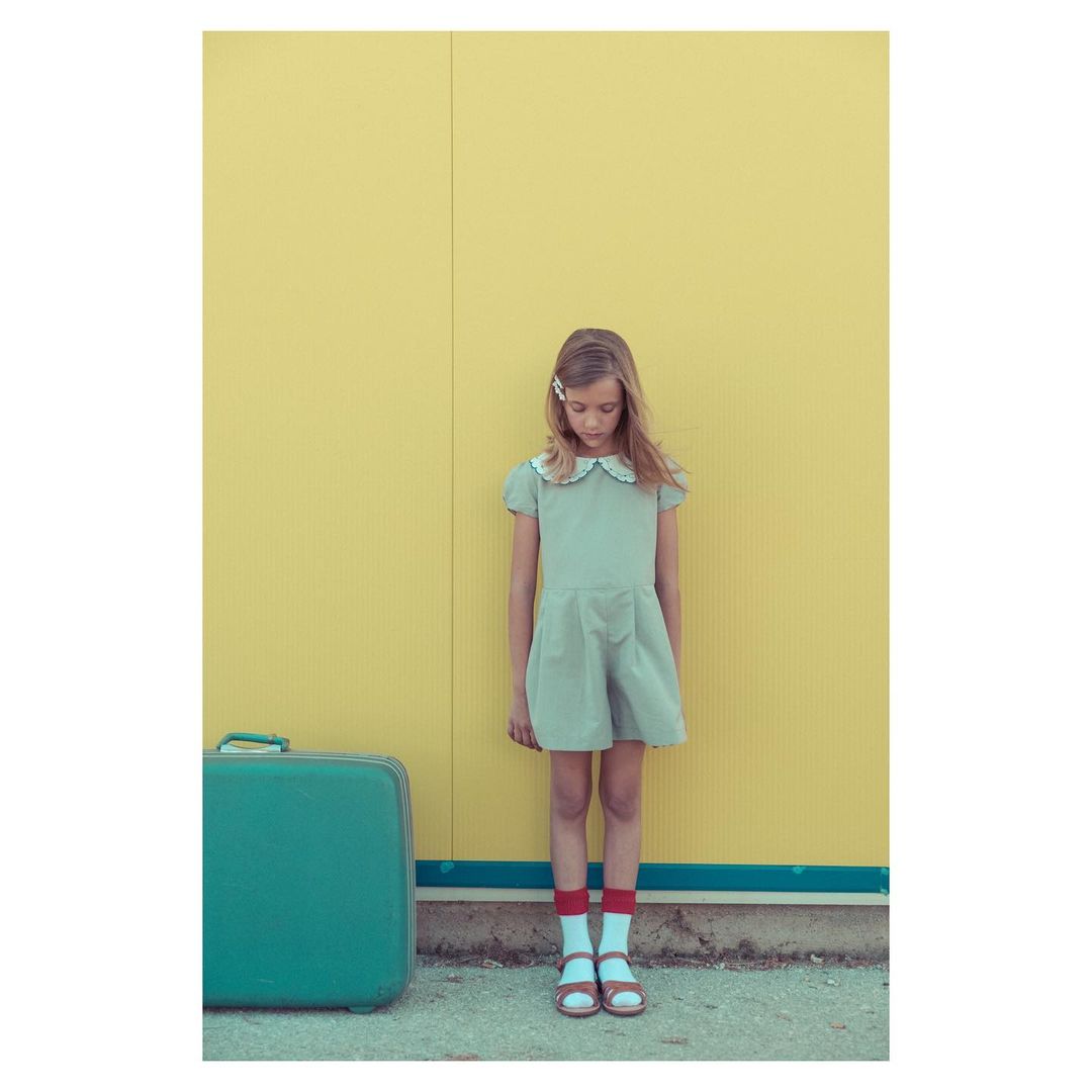Young girl in dress with vintage suitcase stands against yellow wall, wearing red socks and sandals in an outdoor setting.