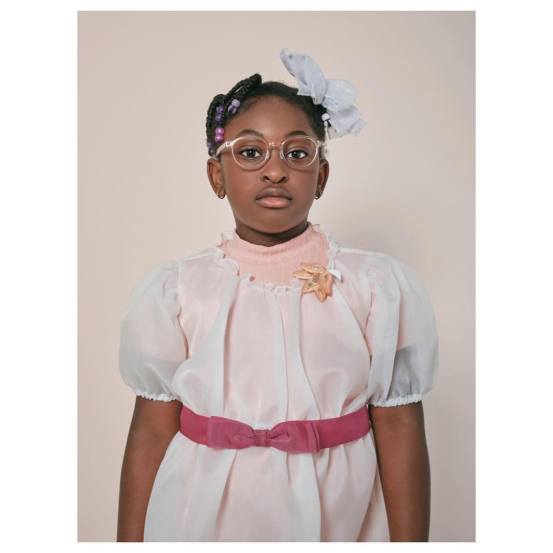 Portrait of a young African-American girl wearing glasses, pink dress with bow, and a decorative hairpiece in a light studio setting.