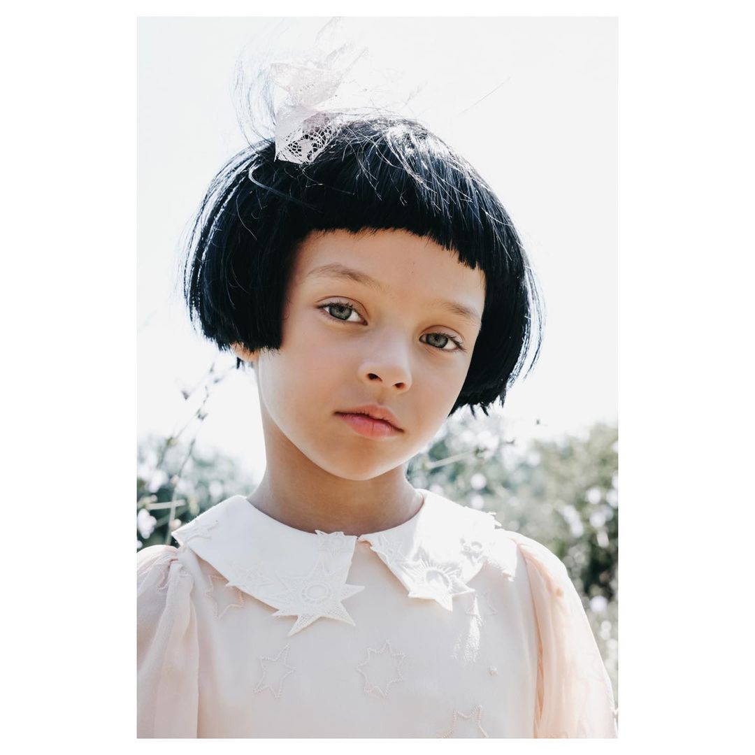 Portrait of a child with bob haircut wearing a white-collar dress with star embellishments, posing outdoors in natural light.
