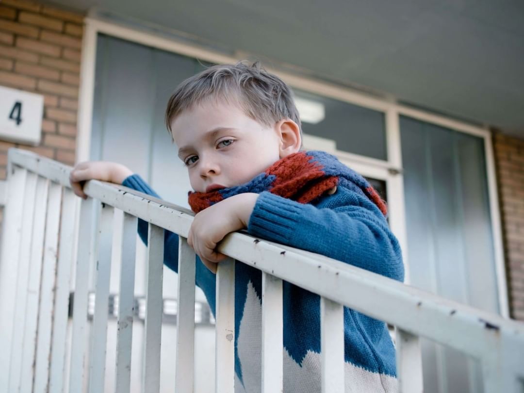 Young boy with a serious expression leans against a white railing outside an apartment building while wearing a blue sweater and scarf.