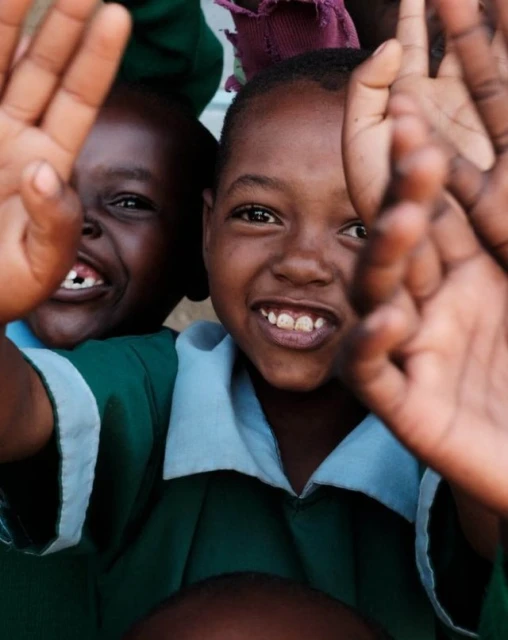 Two smiling Black children in green school uniforms raise their hands to the camera, showcasing their joyful expressions in an outdoor setting.