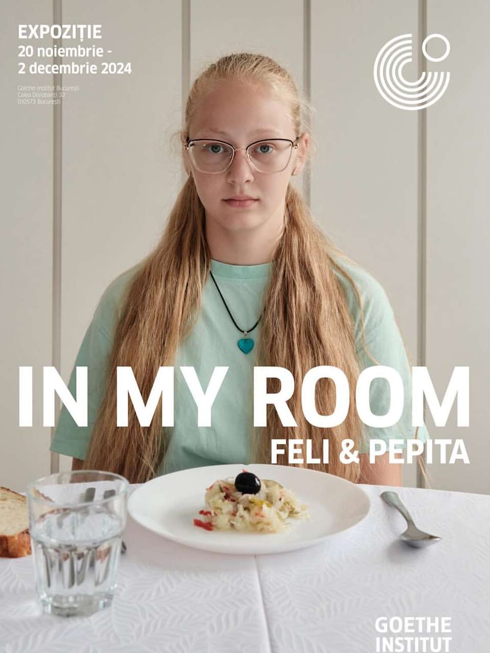 Portrait of a young blonde woman wearing glasses and a teal shirt, seated at a table with a plate of food, in an indoor setting.