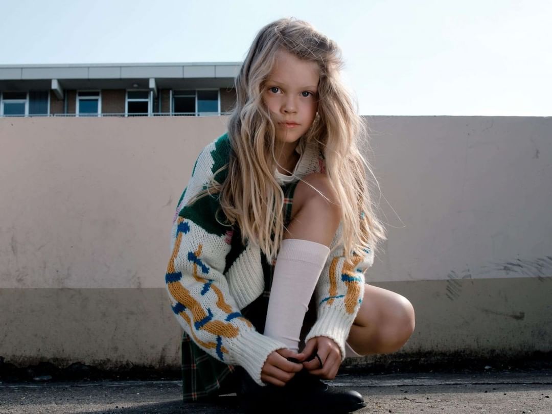 Young girl with long blonde hair, wearing a patterned cardigan and knee socks, crouches outside on a sunny day.