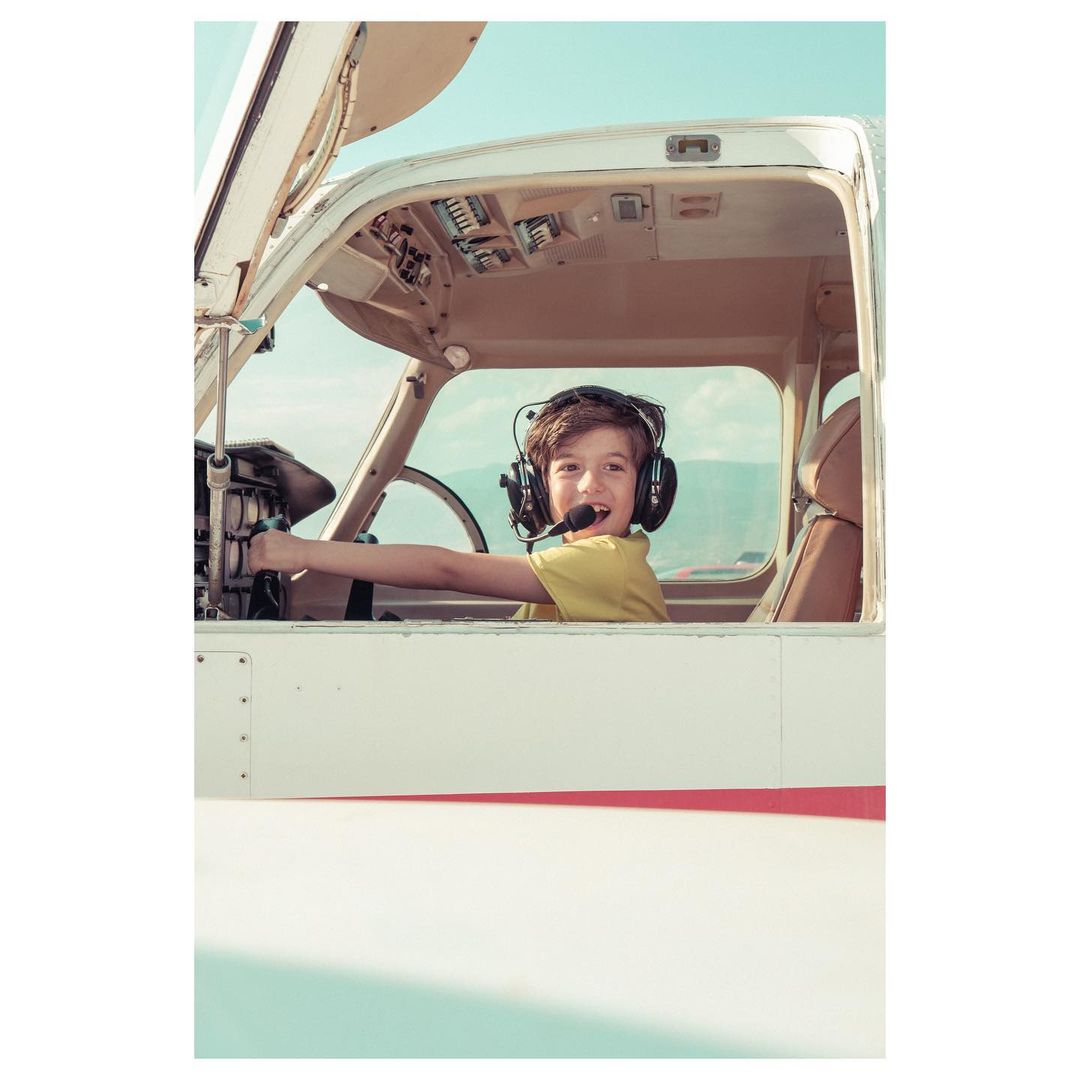 Young boy dressed as a pilot, smiling in the cockpit of a small airplane with an open door, set against a bright blue sky.