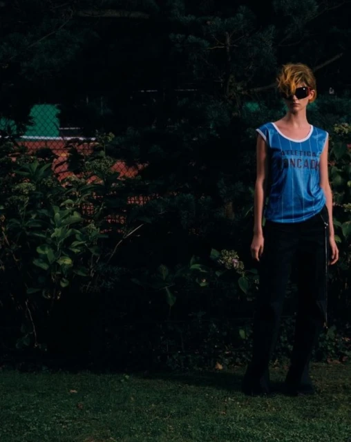 An adult person stands outside wearing a blue tank top and black pants, posing in front of a green lawn and dark hedges.