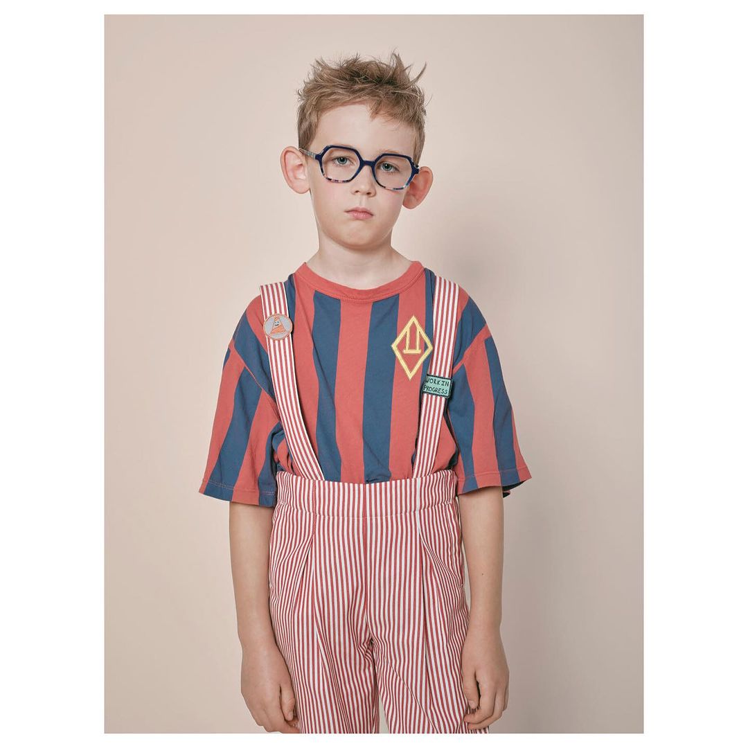 Young boy in eyeglasses and striped outfit poses in a portrait-style shot, showcasing fashion clothing against a neutral background.