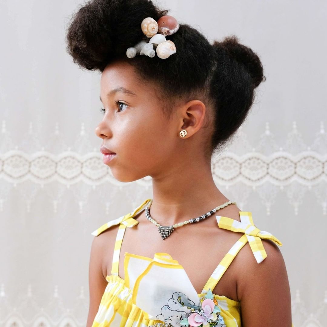 Cute little girl with natural afro hairstyle decorated with seashells, wearing yellow gingham top and a beaded necklace, looking away.
