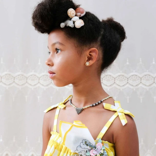 Cute little girl with natural afro hairstyle decorated with seashells, wearing yellow gingham top and a beaded necklace, looking away.