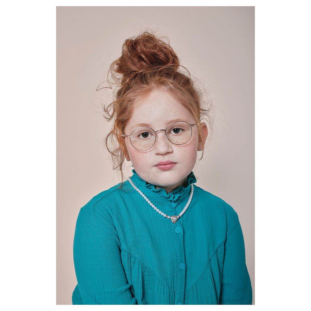 Portrait of a young girl with freckles and red hair in a bun, wearing a teal top and a pearl necklace, set against a neutral backdrop.