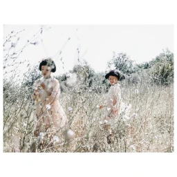 Two children in pale dresses standing in a tall wildflower field under bright daylight by Feli and Pepita