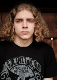 Close-up portrait of a young man with long curly blond hair wearing a black graphic t-shirt against dark wood planks.