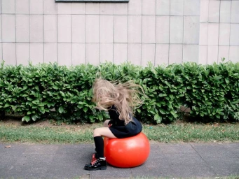 A young girl with long blonde hair bouncing on a bright red hopper ball outdoors against a low green hedge.