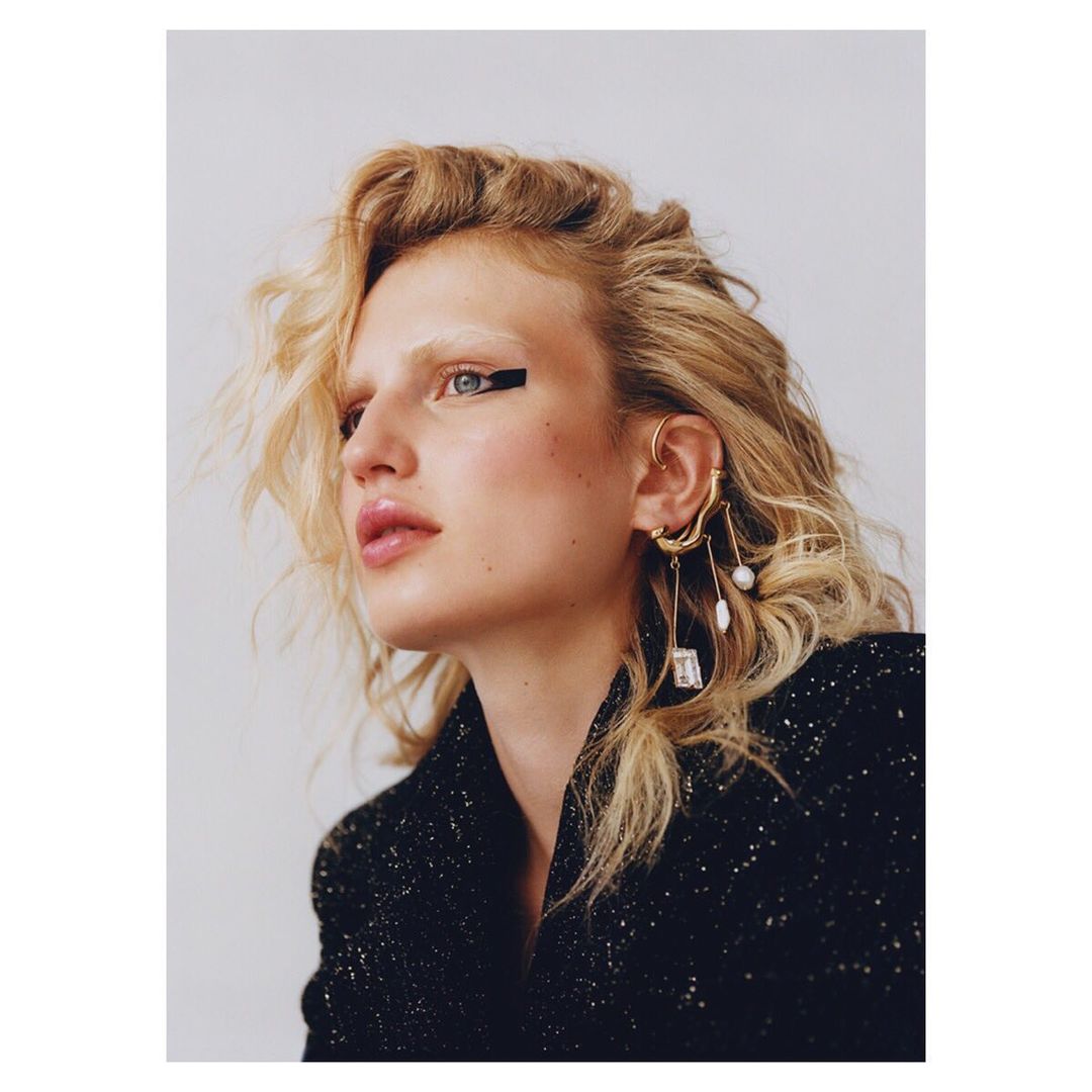Close-up portrait of a woman with blonde wavy hair, wearing statement earrings, a black jacket, and bold black eyeliner.