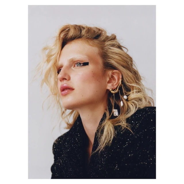 Close-up portrait of a woman with blonde wavy hair, wearing statement earrings, a black jacket, and bold black eyeliner.
