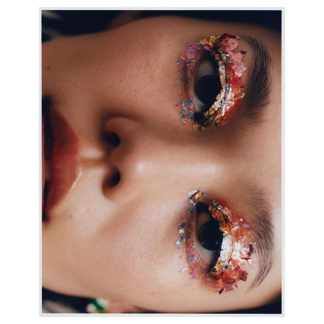 Close-up portrait of a young adult female's face, featuring creative eye makeup with gold, pink, and blue glitter.
