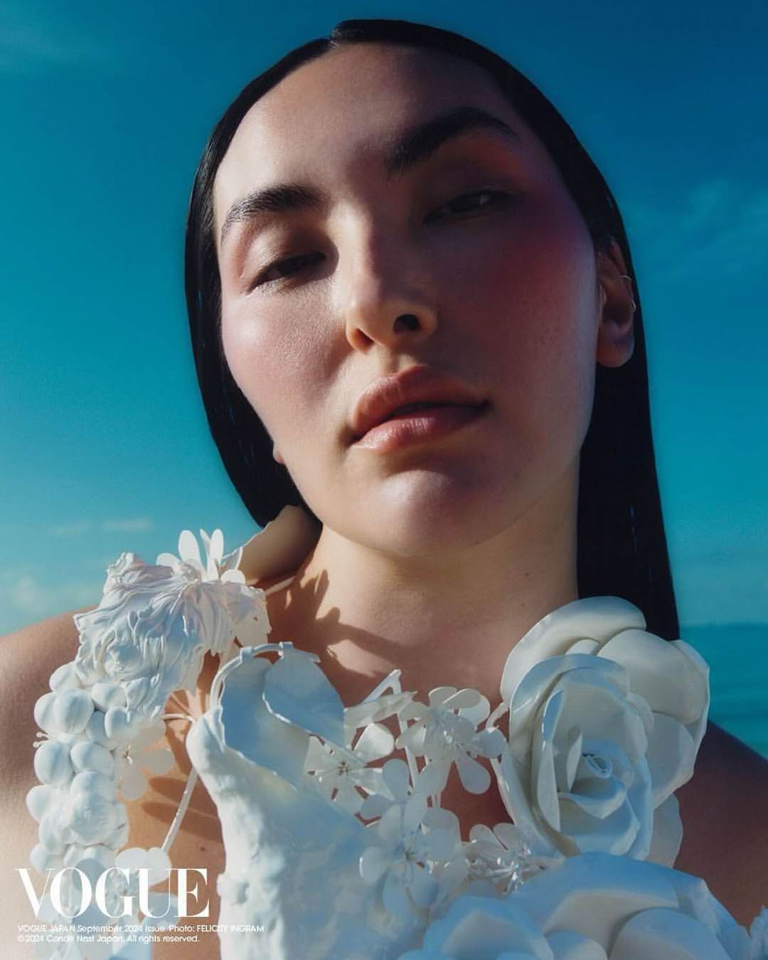Close-up of a woman with long dark hair wearing a white floral garment against a bright blue sky, Vogue logo visible.
