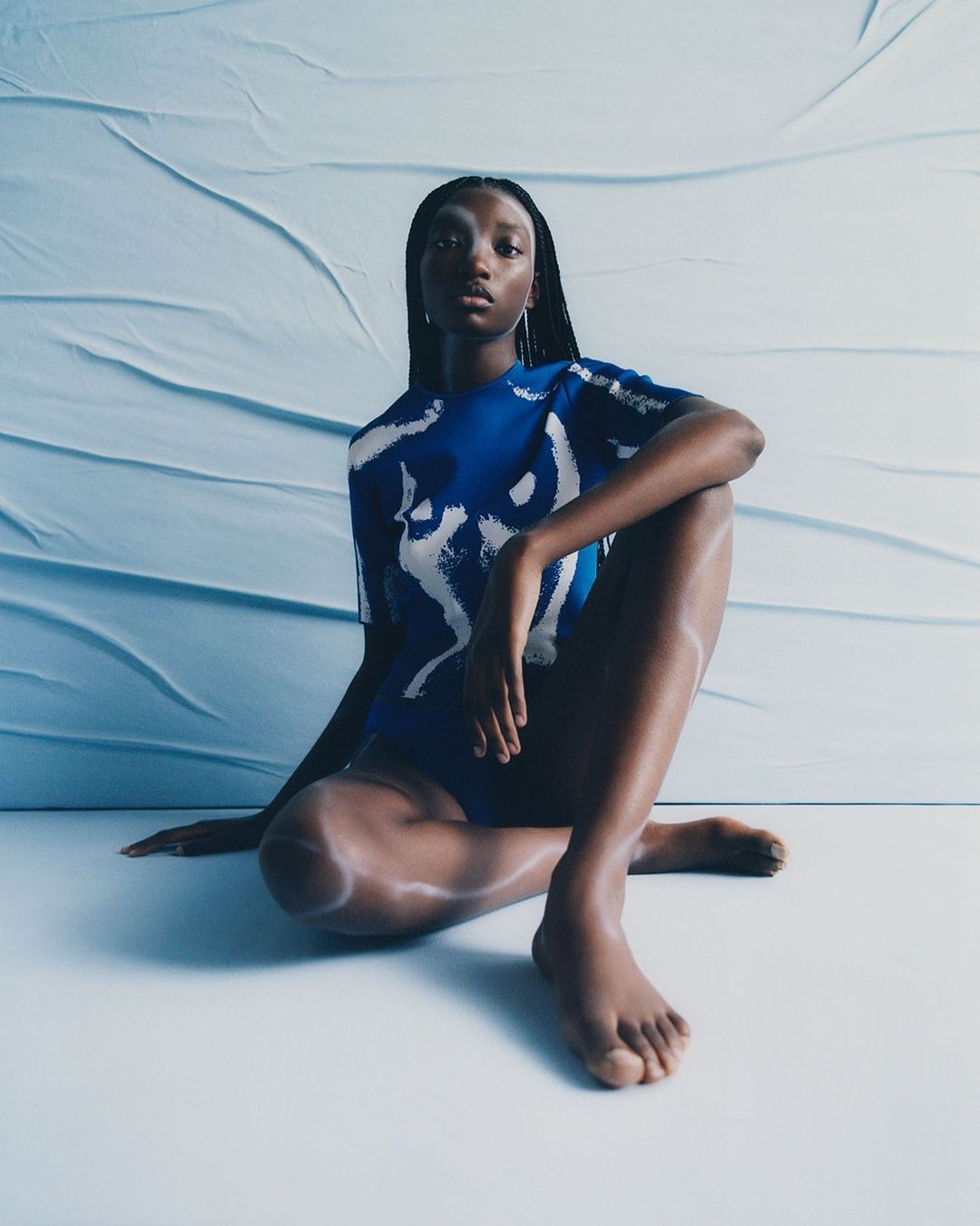 A solo, dark-skinned female model sits cross-legged in a studio, showcasing a blue-and-white patterned bodysuit, with long braided hair and serious expression.