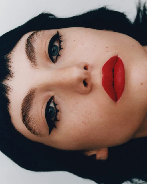 Portrait of a woman with striking makeup featuring black winged eyeliner and red lipstick, set against a simple white background.