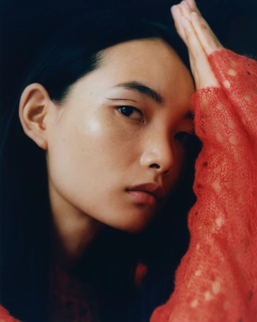 Close-up portrait of a young Asian woman with dark hair and red sweater with hands near her face against a dark background.