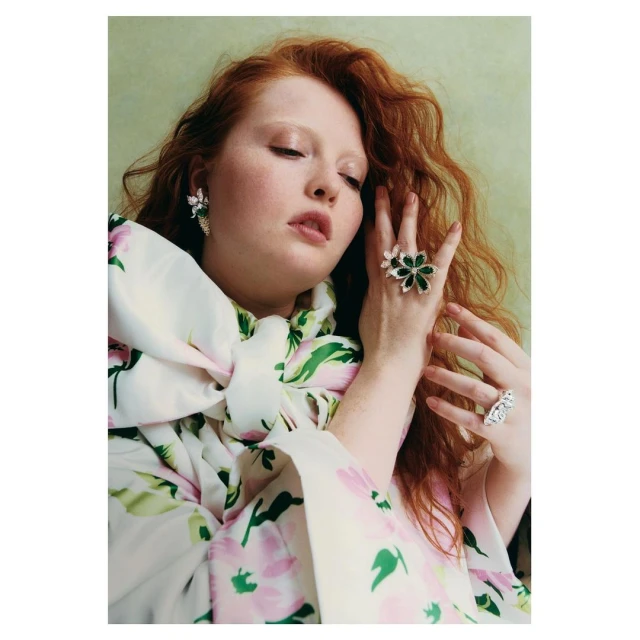 Adult female redhead poses wearing floral dress and floral and gemstone jewelry including earrings and large rings against light-green backdrop.