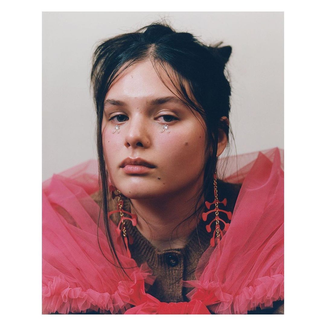 Portrait of a young woman with face gems and pink tulle collar, wearing statement earrings in a brightly lit indoor setting.