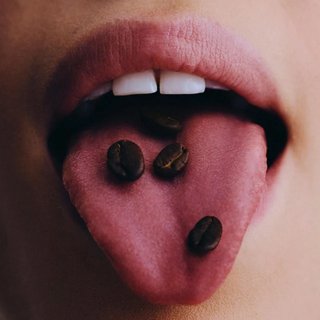 Close-up of a person's mouth with coffee beans on their tongue, showcasing the dark brown color and texture of the beans.