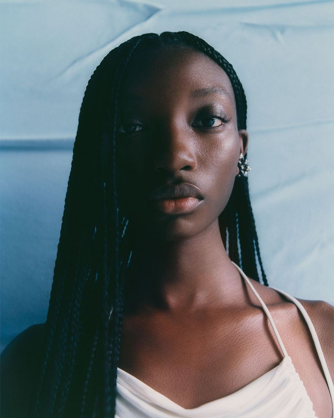 Close-up portrait of a young adult Black woman with braided hair and pale top in front of a pale blue background, looking at the camera.