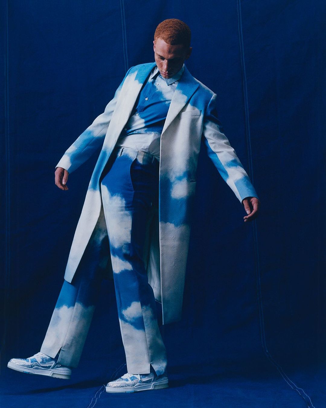 Fashion photo of a red-haired male model in a white coat and cloud-print pants, striking a pose against a dark blue backdrop.