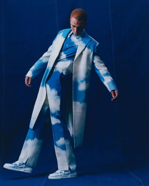 Fashion photo of a red-haired male model in a white coat and cloud-print pants, striking a pose against a dark blue backdrop.