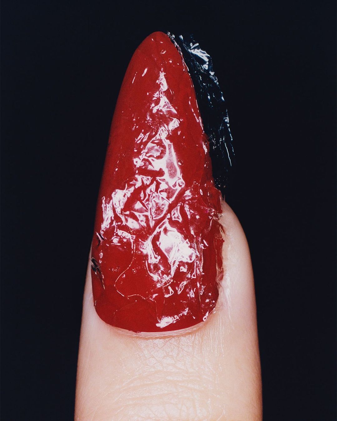 Close-up of a manicured fingernail featuring a glossy red polish and a section of dark foil texture, set against a stark black backdrop.