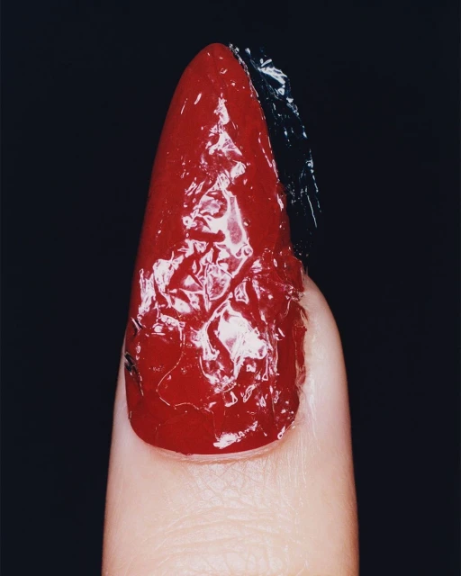Close-up of a manicured fingernail featuring a glossy red polish and a section of dark foil texture, set against a stark black backdrop.