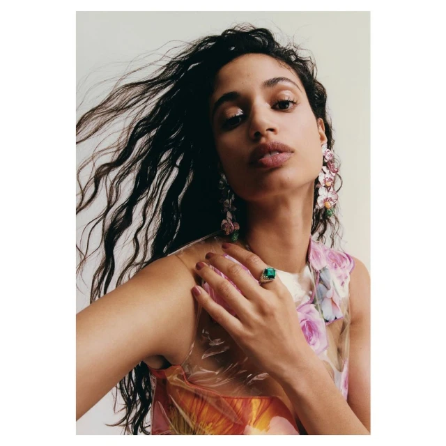 Portrait of a woman with dark, wavy hair wearing a floral dress and flower earrings, showcasing a ring with a green gemstone.