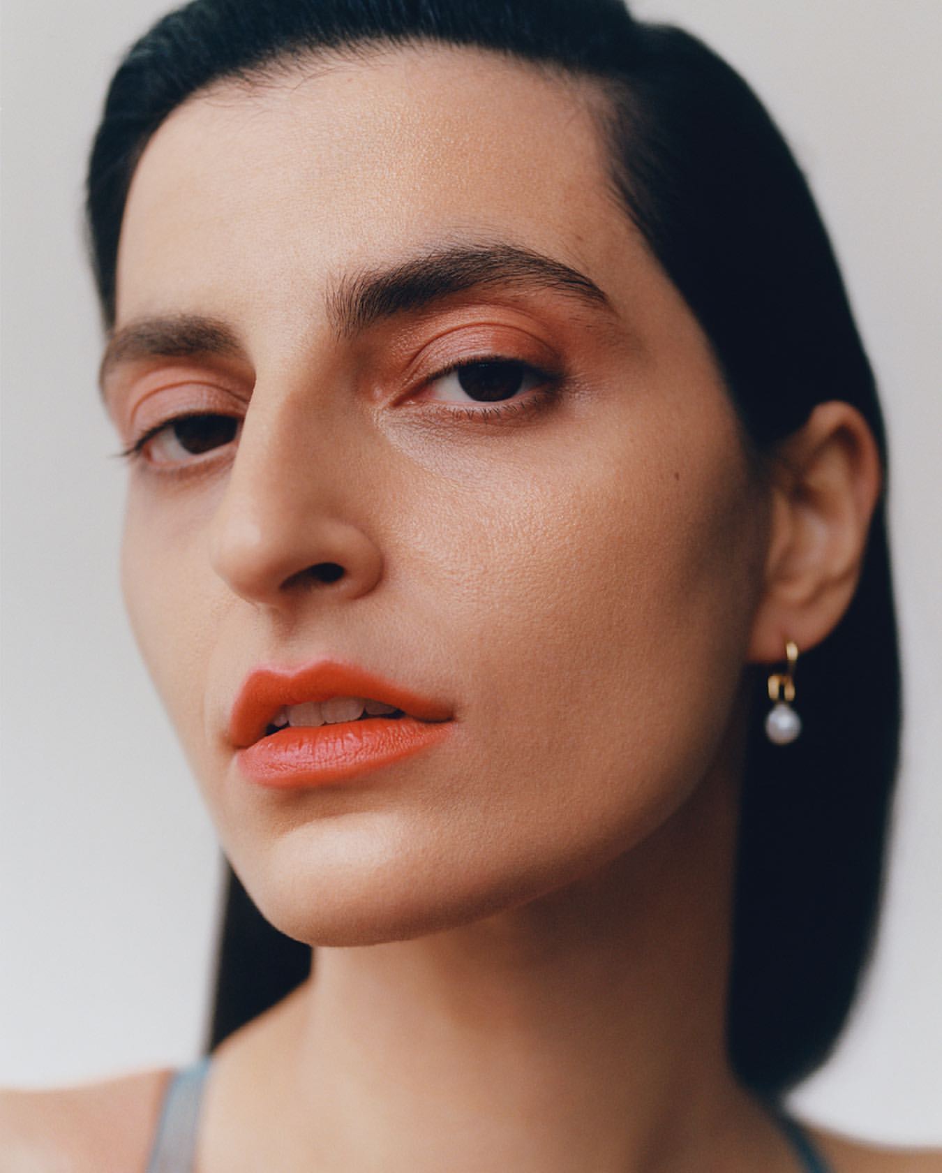 Portrait of a woman with coral lipstick and matching eyeshadow, gold hoop earrings, and black hair, posing against a light background.