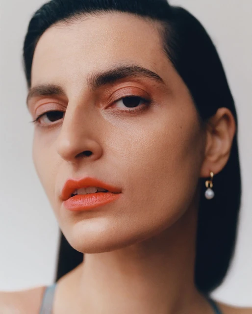Portrait of a woman with coral lipstick and matching eyeshadow, gold hoop earrings, and black hair, posing against a light background.