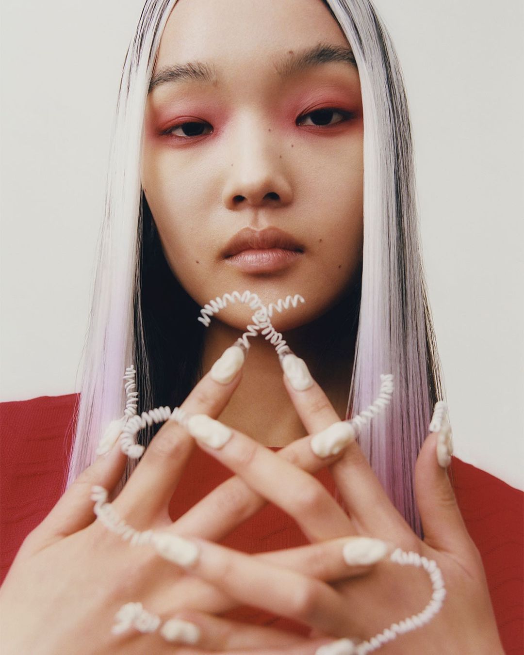Close-up of Asian woman with white hair, striking red eyeshadow, and sculptural white nail art, posing in front of a plain background.