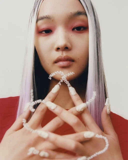 Close-up of Asian woman with white hair, striking red eyeshadow, and sculptural white nail art, posing in front of a plain background.