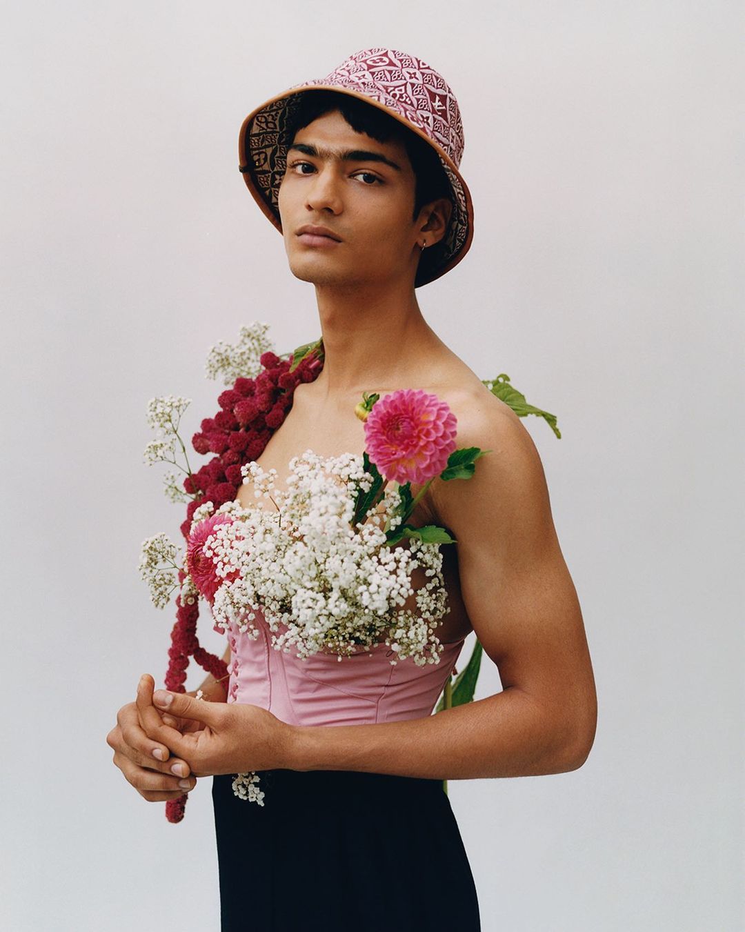 Portrait of a person wearing a pink top and bucket hat, adorned with flowers, standing against a plain background, looking at camera.