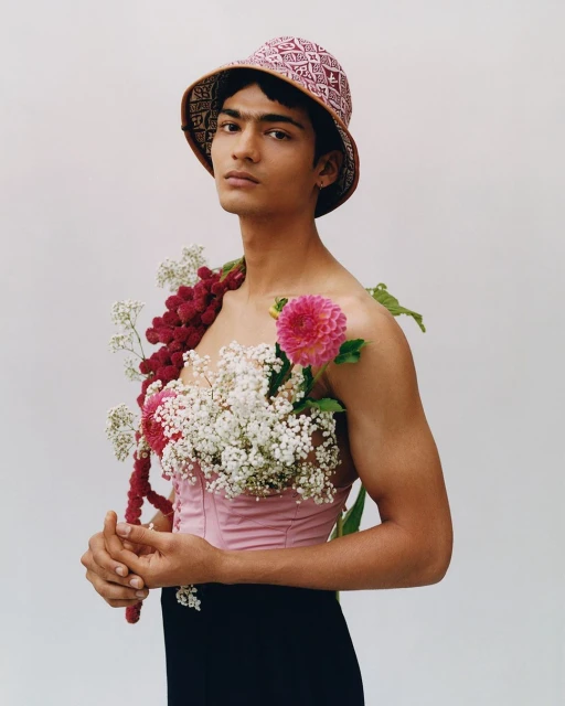 Portrait of a person wearing a pink top and bucket hat, adorned with flowers, standing against a plain background, looking at camera.