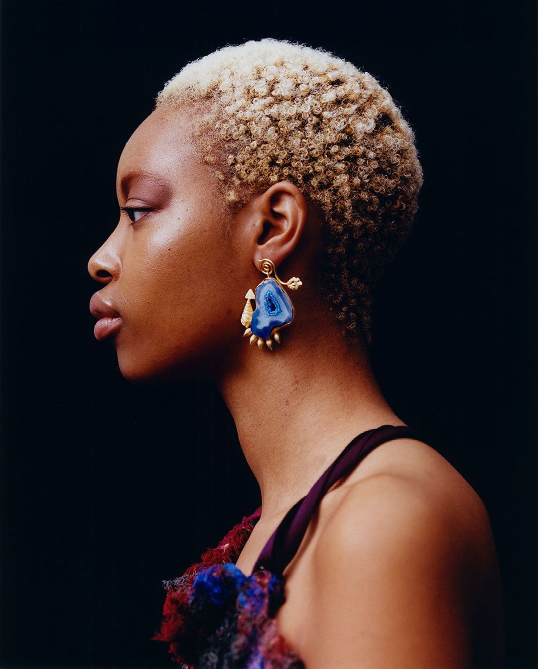 Profile shot of an adult female with blonde textured hair and blue stone earrings against a dark background in a studio setting.