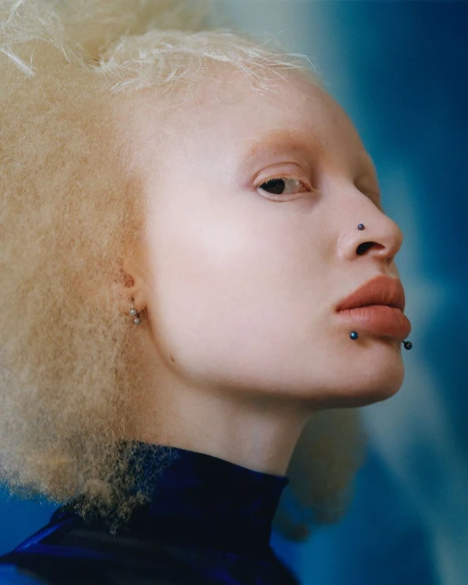 Close-up studio portrait of an adult female with fair skin, light blonde afro-textured hair, and dark blue top against a blue background.