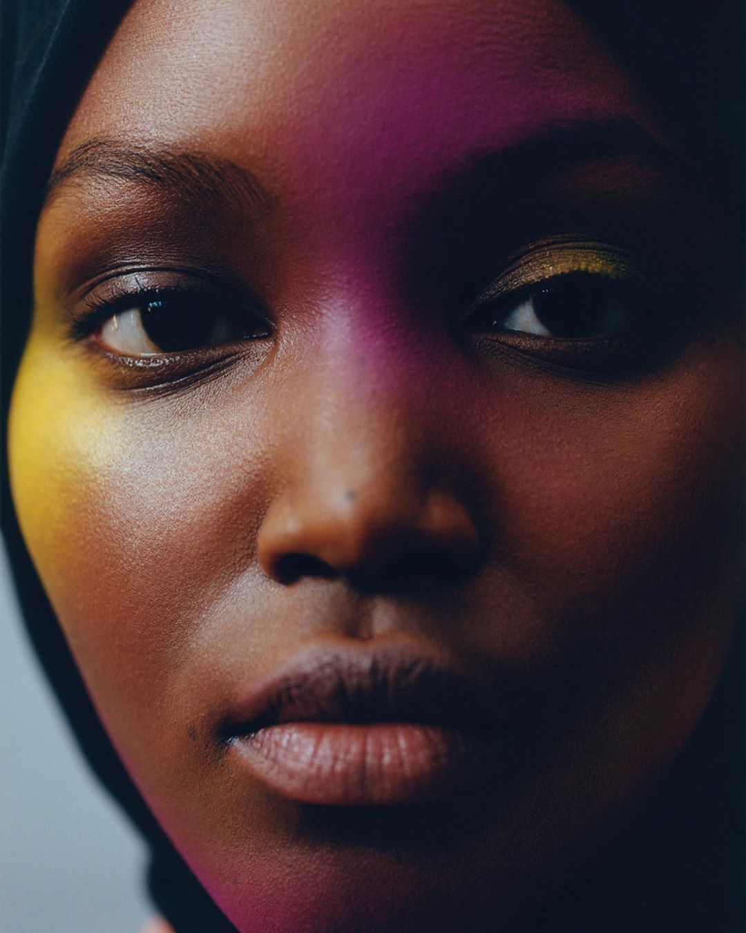 Close-up portrait of a Black woman with colorful lighting, wearing a dark head covering, showcasing beauty and skincare.